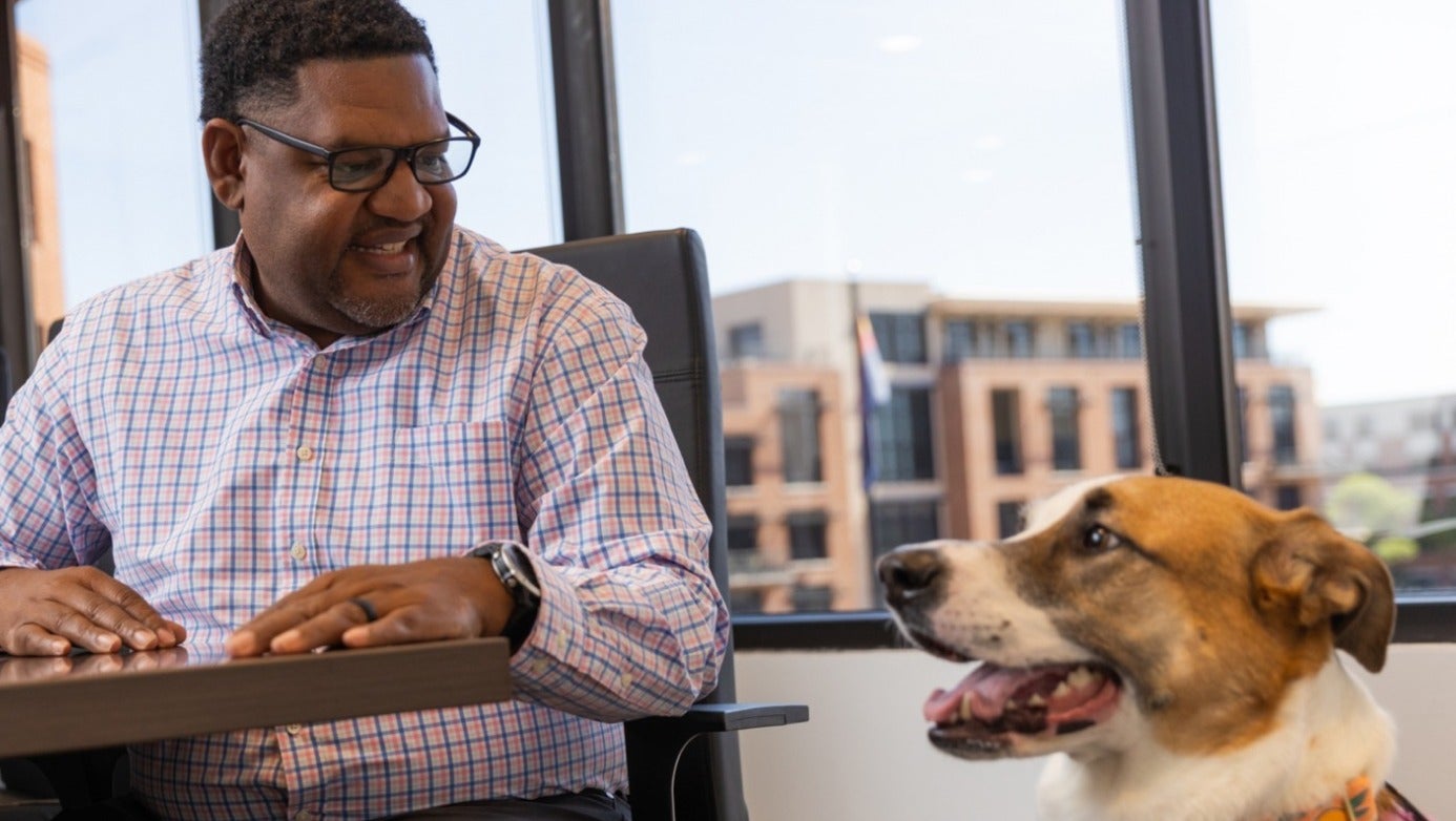 A man wearing glasses sits at a table, enjoying the company of a dog beside him.