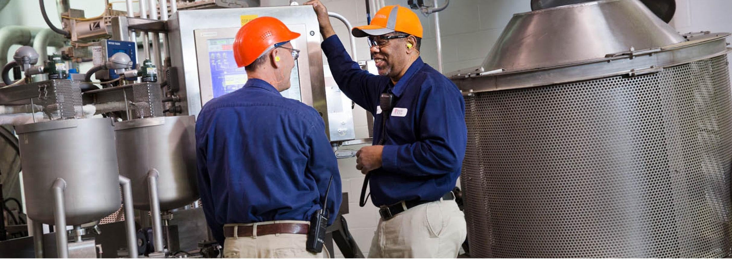 Two men in blue shirts and hats stand before a large industrial machine, showcasing teamwork in a manufacturing setting.