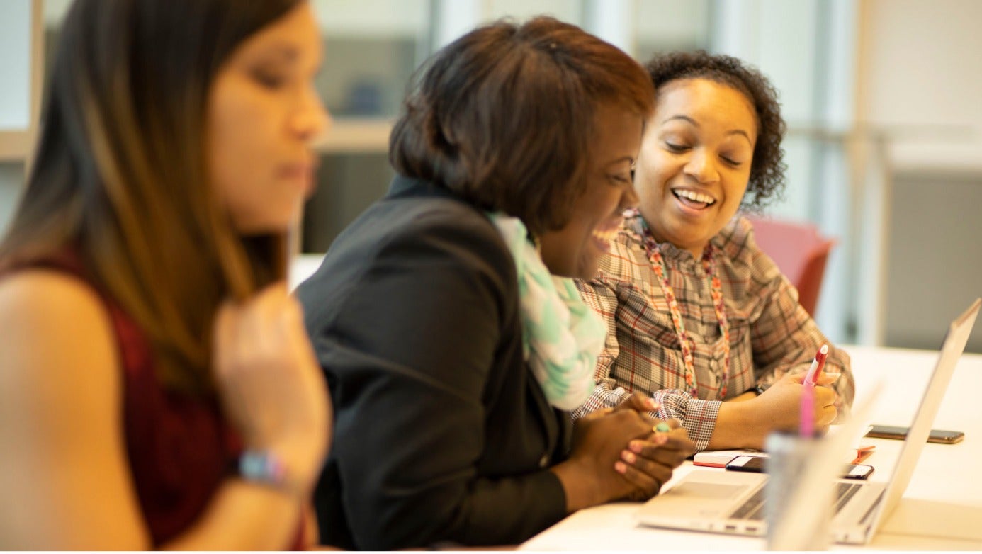 Three women engaged in work at a table, each using a laptop, showcasing collaboration and productivity.