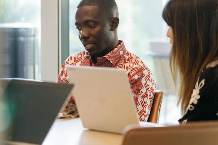 Two employees look at computer in shared discussion. Two open computer are on display on flat top table. 